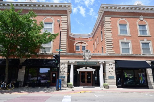 Historic brick building with ornate details, featuring large entrance doors and shops on either side under a blue sky.