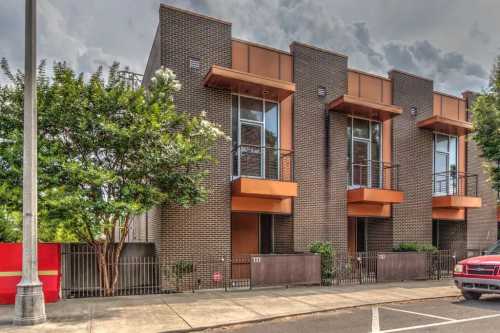 Modern brick townhouses with large windows and balconies, surrounded by greenery and a sidewalk. Cloudy sky above.