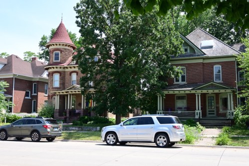 Two historic houses with distinct architectural styles, surrounded by trees, and parked cars on a sunny street.