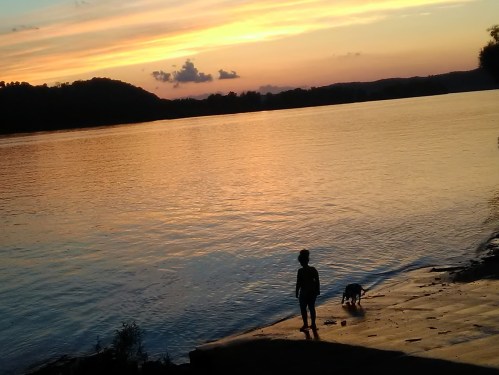A silhouette of a child and a dog by the water at sunset, with colorful skies and hills in the background.