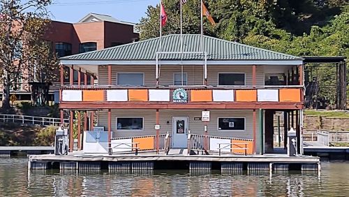 A two-story marina building with orange accents, flags on the roof, and a dock on a calm waterway.