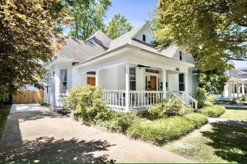 A charming white house with a porch, surrounded by greenery and trees on a sunny day.