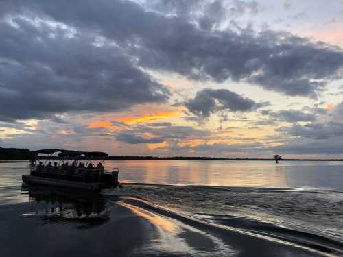 A boat with passengers glides across a calm lake at sunset, with colorful clouds reflecting on the water.