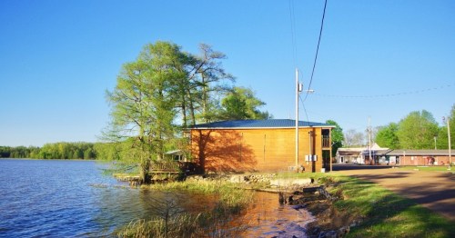 A wooden building by a calm lake, surrounded by trees and a clear blue sky, with a path along the water's edge.