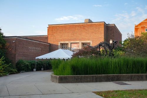 A brick building with a tent in front, surrounded by greenery and a sculpture of a dinosaur.