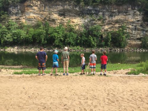 Six children stand by a riverbank, facing a rocky cliff, with greenery surrounding them on a sunny day.