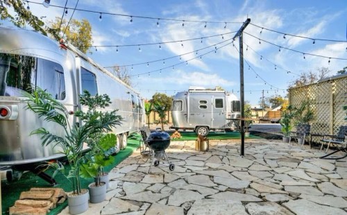 A cozy outdoor space featuring two silver Airstream trailers, string lights, and a stone patio with plants and a grill.
