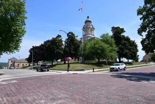 A historic building with a dome stands on a green hill, surrounded by trees and a brick-paved street under a clear blue sky.