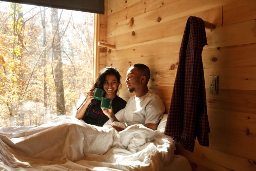 A couple sits in bed, smiling and holding mugs, with sunlight streaming through a window in a cozy wooden cabin.