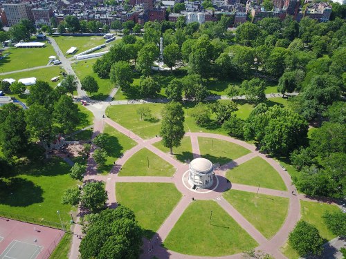 Aerial view of a park featuring a circular gazebo surrounded by pathways and lush greenery. City buildings in the background.