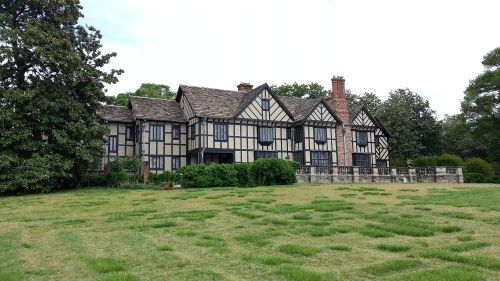 A large, traditional Tudor-style house with black and white timber framing, surrounded by a grassy lawn and trees.