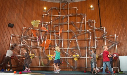 Children interact with a colorful, large wall maze made of tubes, exploring and playing with balls.