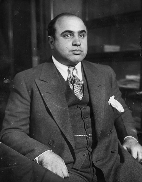A man in a suit sits confidently, looking directly at the camera, with a serious expression and a handkerchief in his pocket.