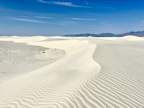 A vast desert landscape with rolling white sand dunes under a clear blue sky and distant mountains.