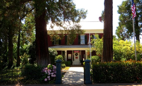 A red two-story house surrounded by greenery and flowers, with an American flag and a welcoming sign on the porch.