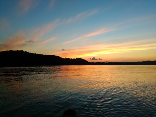 A serene sunset over calm waters, with colorful clouds reflecting on the surface and hills in the background.