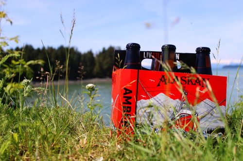 A six-pack of Alaskan beer sits in tall grass by a lake, surrounded by trees and a clear blue sky.