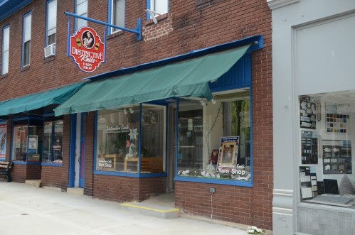 A brick storefront with a green awning, featuring a sign for "Distinctive Knits Yarn Shop" and a display window.