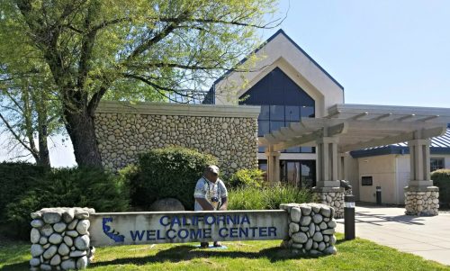 A stone building with a sign reading "California Welcome Center" and a statue in front, surrounded by greenery.