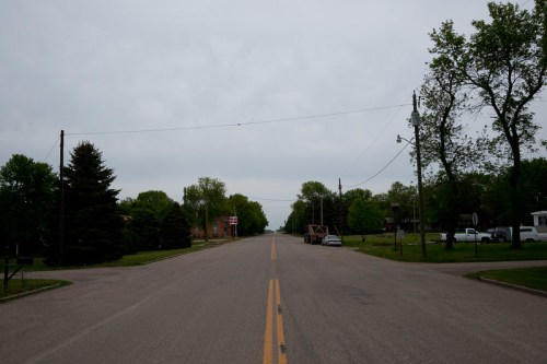 A quiet, empty road lined with trees and houses under a cloudy sky. Power lines stretch across the scene.