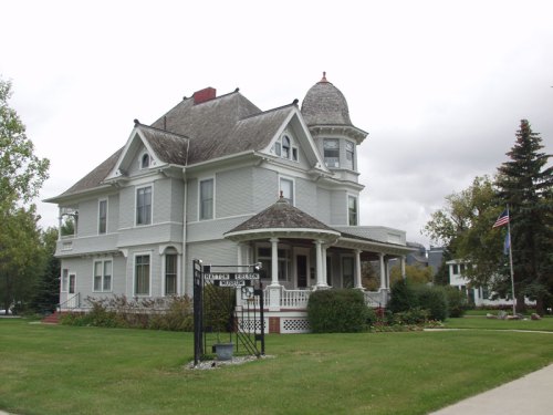 A large, historic gray Victorian house with a turret, surrounded by green grass and trees, under a cloudy sky.