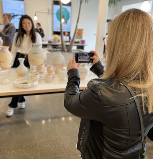 A woman takes a photo of another woman smiling in a gallery filled with colorful pottery and art.