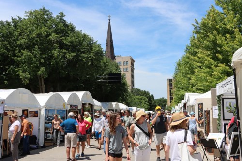 A bustling outdoor art fair with people browsing booths, trees lining the street, and a church spire in the background.