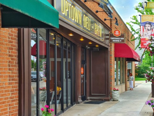 Exterior view of Uptown Deli and Brew, featuring large windows and a brick facade, with nearby shops and flowers.
