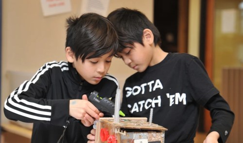 Two boys work together on a craft project, using a glue gun on a box made of various materials.