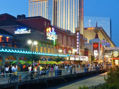 A lively boardwalk scene at dusk, featuring colorful lights, crowds, and nearby tall buildings.