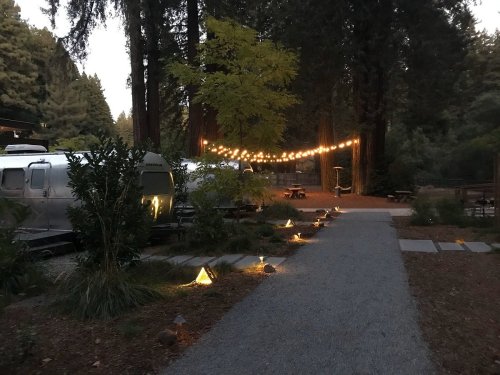 A serene campsite at dusk, featuring string lights, Airstream trailers, and a gravel path surrounded by trees.