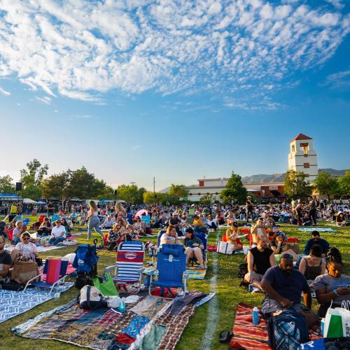 A large crowd gathers on a grassy field with blankets and chairs, enjoying a sunny day under a blue sky with clouds.