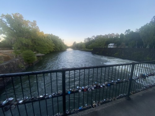 A serene river flows between lush green banks, with a bridge adorned with padlocks in the foreground.