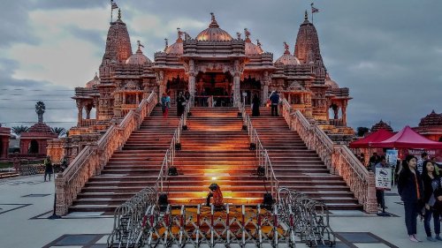 A grand temple with intricate architecture, featuring steps leading up to the entrance, surrounded by visitors and cloudy skies.