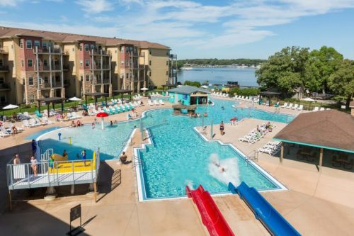 A vibrant pool area with water slides, lounge chairs, and a scenic view of the lake and buildings in the background.
