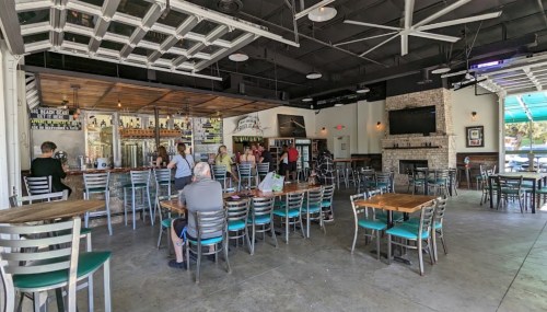 A spacious restaurant interior with wooden tables, metal chairs, a bar area, and patrons enjoying their meals.