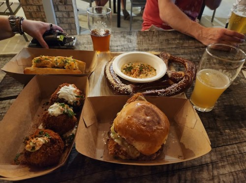 A table with various dishes: a burger, pretzel, loaded fries, and croquettes, alongside two glasses of beer.
