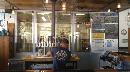 A bartender serves drinks at a brewery, with taps and a colorful menu board in the background.
