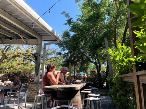 A sunny outdoor bar scene with patrons sitting at a wooden table surrounded by trees and greenery.