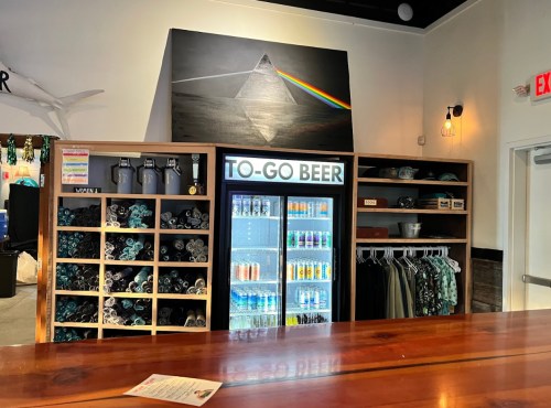 A bar area featuring shelves of merchandise, a fridge labeled "TO-GO BEER," and a colorful pyramid artwork above.