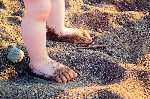 A child's feet covered in sand, standing on a sandy beach with small stones scattered around.