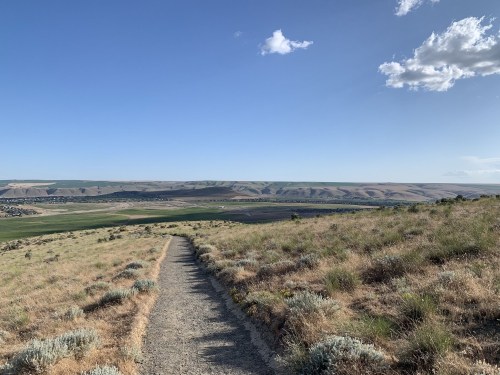 A winding dirt path leads through grassy hills, with a vast valley and blue sky in the background.