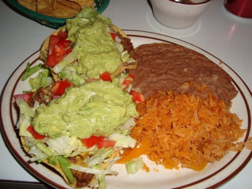 A plate of tacos topped with lettuce, tomatoes, and guacamole, served with refried beans and orange rice.