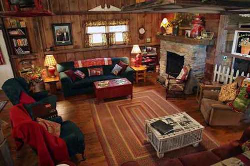 Cozy rustic living room with wooden walls, a stone fireplace, comfortable seating, and warm lighting.