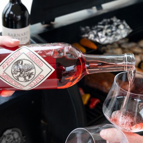 A bottle of rosé wine being poured into a glass, with a grill and food in the background.