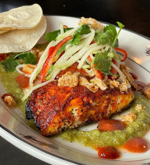 A plate of grilled fish topped with shredded vegetables, herbs, and crispy toppings, served with tortillas and sauce.