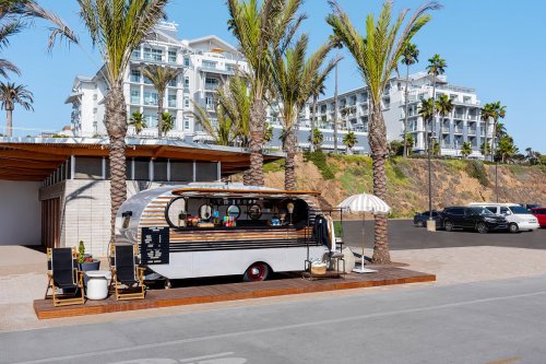 A stylish food truck parked by palm trees, with a modern building in the background and outdoor seating nearby.