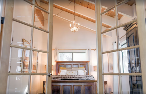 A cozy bedroom with a wooden bed, chandelier, and large windows, viewed through glass-paneled doors.