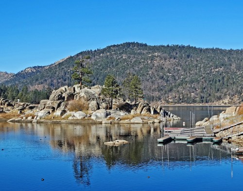 A serene lake scene with rocky shores, pine trees, and a clear blue sky reflecting on the water's surface.