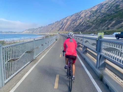 A cyclist rides along a coastal path with mountains and the ocean in the background on a sunny day.